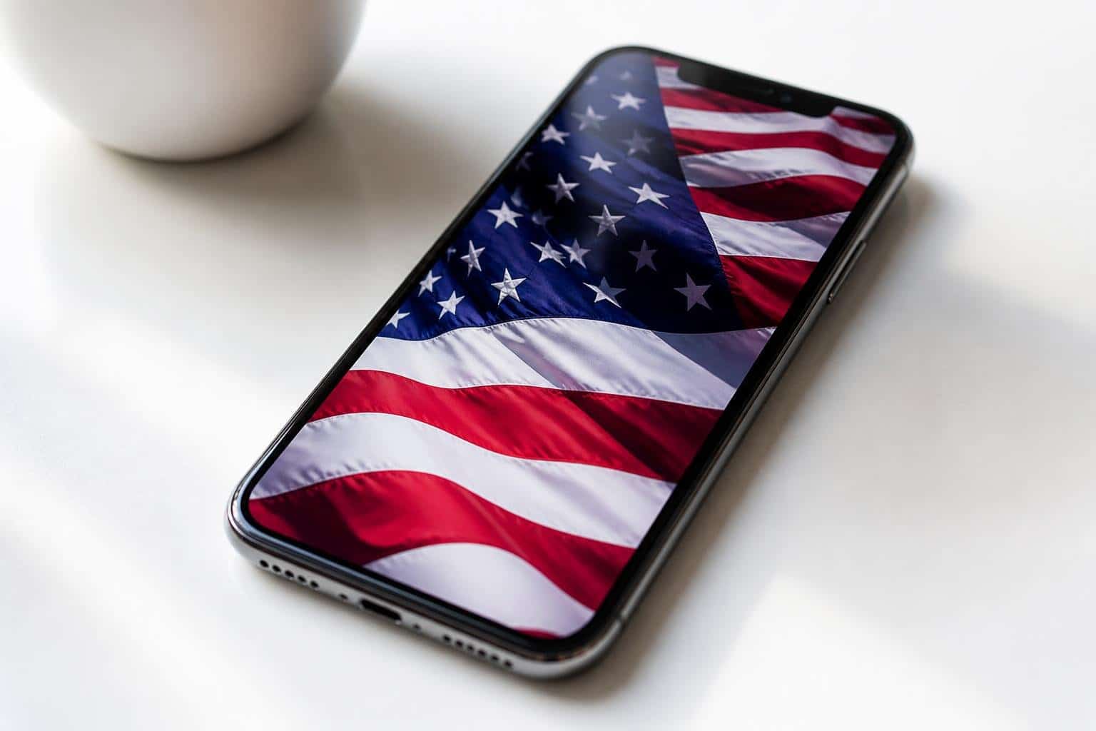 Um iPhone moderno sobre uma superfície branca exibindo a bandeira americana na tela.